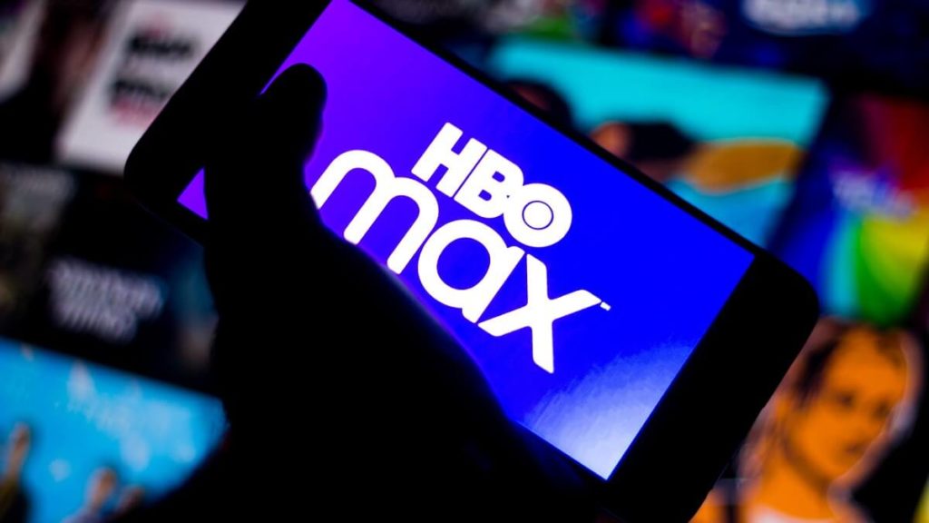 HBO Max na tela de um celular sendo segurado por uma mão em um local escuro com algumas séries de fundo.